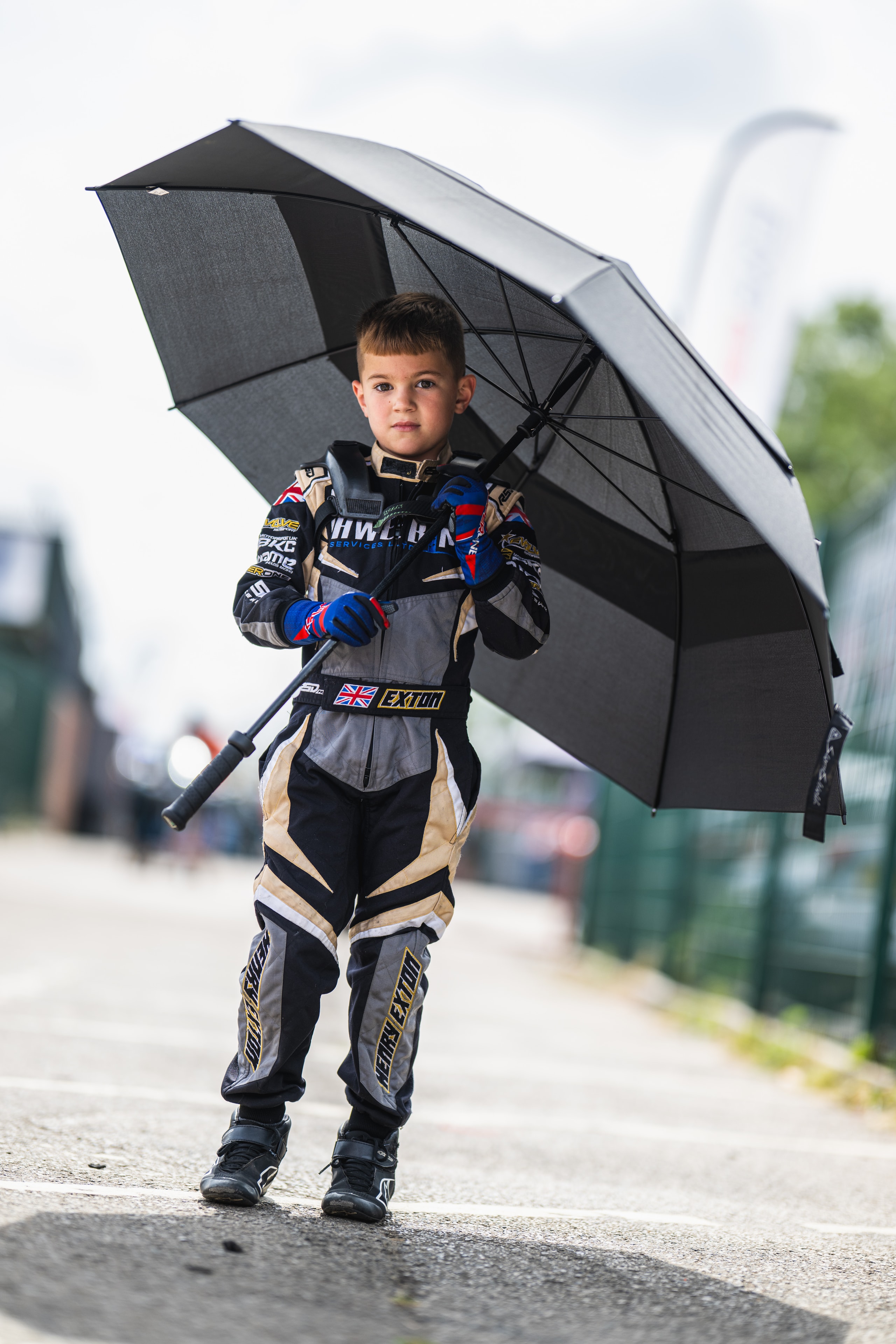 Henry Exton in race suit holding umbrella in the paddock at Whilton Mill